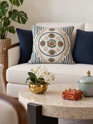 The Noorani embroidered cushion cover on a cream sofa between navy cushions, beside a brass bowl of white orchids on a stone coffee table