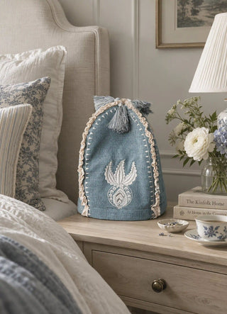 The Noush tea cosy in Dusty Blue, view-bedside beside stacked books and white ranunculus.