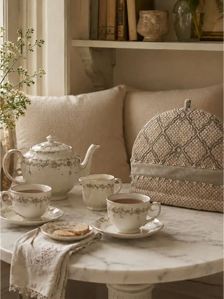 The Oat Pearl insulated tea cozy adding a serene neutral texture to a marble bistro table set with delicate floral china and linen napkins.