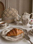 The Oat Pearl insulated tea cosy styled on a white tablecloth next to a vintage sepia porcelain teapot and a fresh croissant.