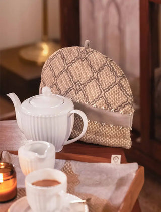 The Oat Pearl tea cosy with a grey lattice motif, pearl-like beads and a soft grey band on a chunky oat weave, with a white teapot on a wooden table.