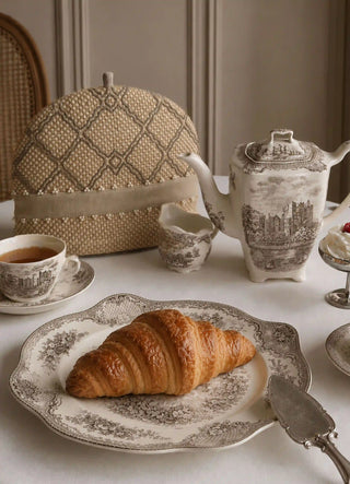 Oat Pearl tea cosy on a marble table beside a vintage floral teapot, cups and a croissant