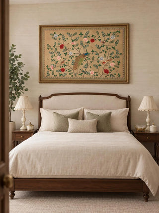 The Pakhi wall art in a gilt frame above an upholstered bed with sage cushions, scalloped lamp shades and a potted tree.