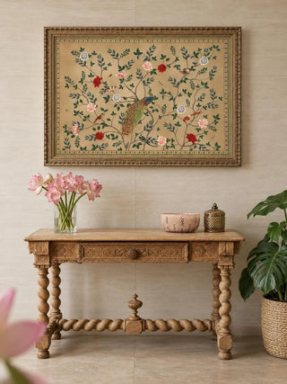 The Pakhi wall art in a carved gilt frame above a barley-twist wooden console with pink flowers in a glass vase and a painted bowl.