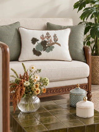 The Palm embroidered rectangular cushion cover on a cream sofa between sage green cushions, beside a glass vase of wildflowers and a green tiled table