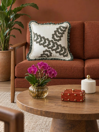 Palmora cushion cover in ivory with sage and taupe scalloped embroidery and a sage ruffle, on a rust-orange sofa with oak arms, beside a round wood coffee table with a pink orchid in a ribbed amber glass vase.