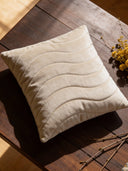 A luxury Pearl Drift quilted cushion cover in champagne velvet with wave-patterned stitching resting on a dark wooden table.