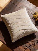 A luxury Pearl Drift quilted cushion cover in champagne velvet with wave-patterned stitching resting on a dark wooden table.