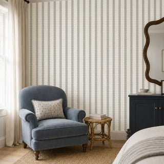 The Pebble Stripe cream grey wallpaper installed behind a blue armchair and dark wood cabinet with a wavy mirror view-bedroom