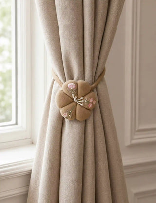 The Petal Knot embroidered puff blossom tie back gathering a heavy oatmeal linen curtain panel next to a white window frame.