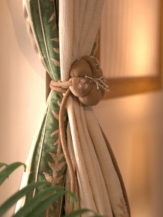 The Petal Knot tie back in tan with a padded knot, pink blossom details and pearl-like beaded sprays, holding a cream and green curtain.