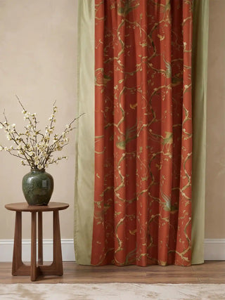 A full-length Phoenix Chinoiserie curtain in terracotta and sage styled next to a wooden side table and floral vase.