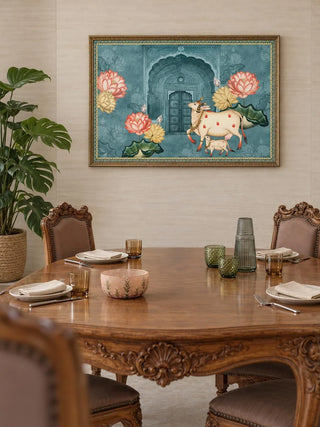 The Pichwai Cow Wall Art in a gilded frame on a neutral wall above a carved round wooden dining table with upholstered chairs.