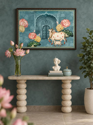 The Pichwai Cow Wall Art in a dark wood frame on a teal wall above a stone console with stacked round legs and a vase of pink lotuses.