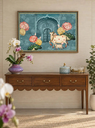 The Pichwai Cow Wall Art in a gilded frame above a scalloped wooden console with brass pulls, a purple vase of orchids and a crystal bowl.