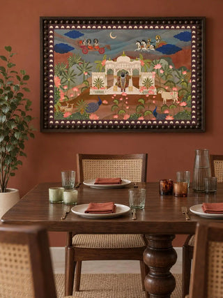 Pichwai Palace embroidered wall art in a dark beaded frame on a terracotta wall above a wooden dining table with cane-back chairs in a dining room