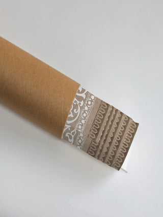 The Pietra wall art print partly unrolled from its shipping tube, showing white scrollwork beside a tan leaf-pattern border.