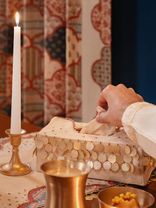 The Pitara tissue box cover in cream with rows of hanging gold discs and scattered colourful beads, with a tissue being drawn from the top.