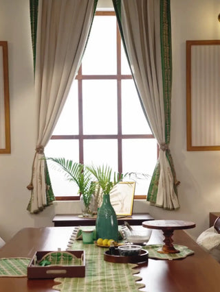Elfdale readymade curtains in beige with a green leaf-sprig border, framing a window over a dining table as a woman draws one open