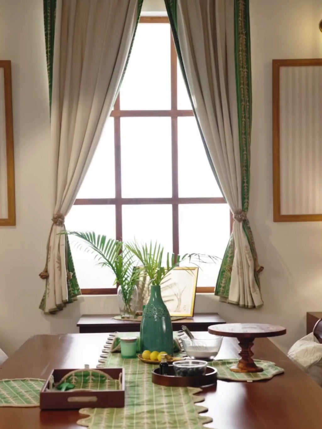 Elfdale readymade curtains in beige with a green leaf-sprig border, framing a window over a dining table as a woman draws one open