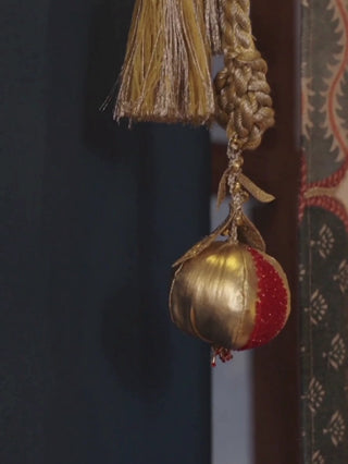 Nar curtain tieback with a gold and red beaded pomegranate, twisted gold cord and tassel, hanging beside a curtain on a blue wall