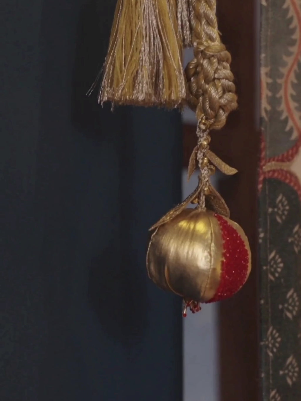 Nar curtain tieback with a gold and red beaded pomegranate, twisted gold cord and tassel, hanging beside a curtain on a blue wall