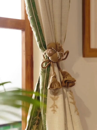 Petal Knot curtain tieback with padded blossoms and rose tassels, gathering a beige curtain with a green border beside a lit lamp