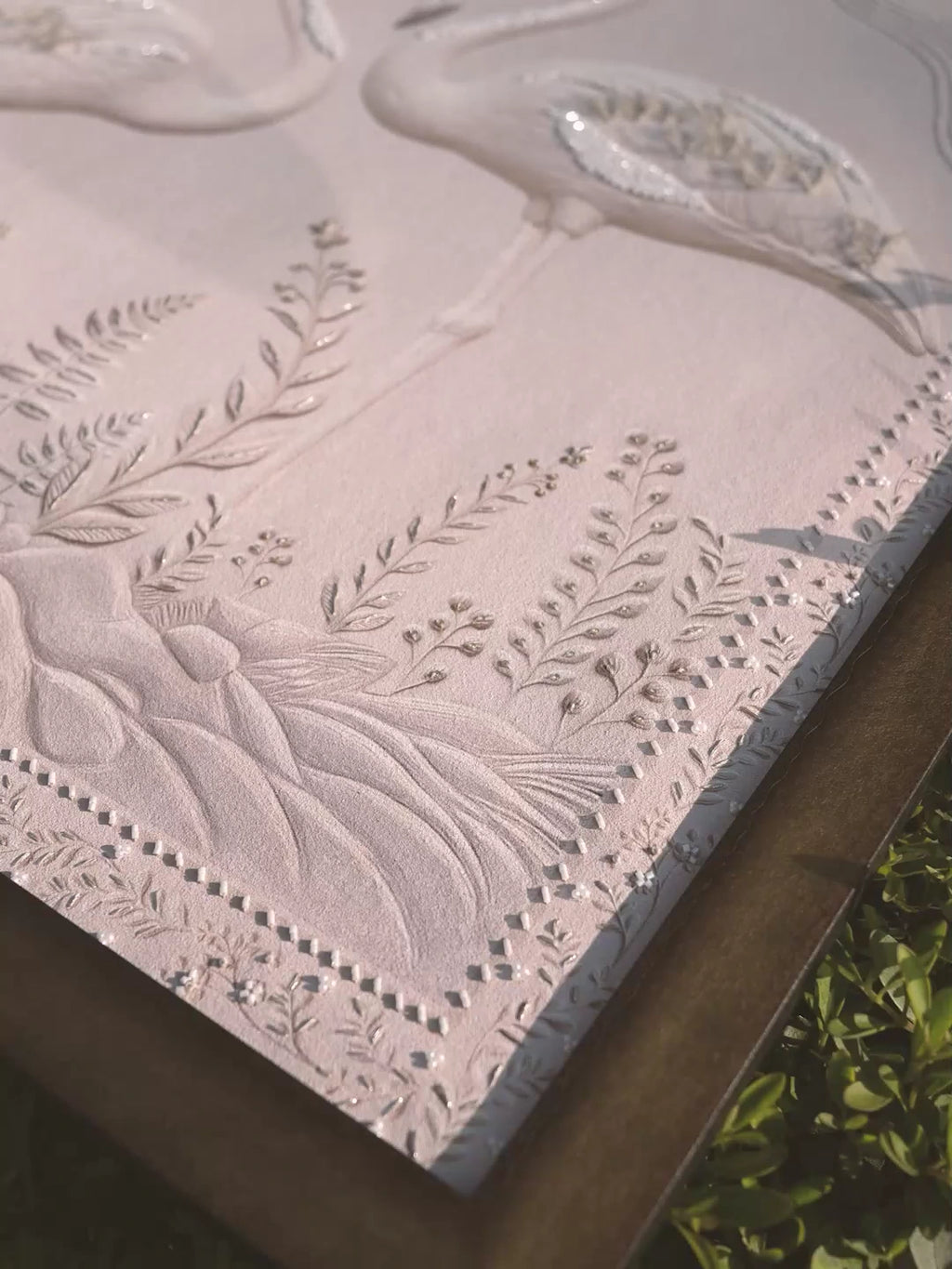 Sculpted Song embroidered wall art in white relief: a flamingo, ferns and sequins raised on a pale ground, filmed at an angle outdoors