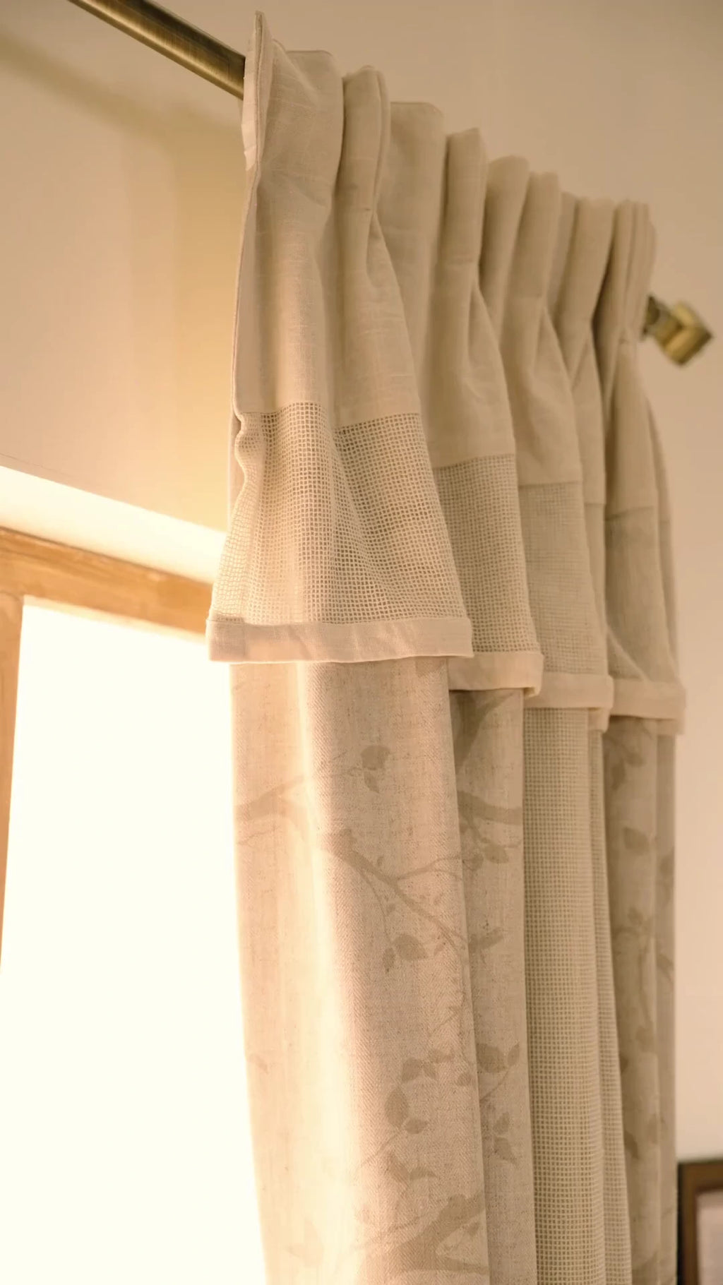 The Reed Rhythm readymade curtain in beige with a faint botanical print, shown in pleats on a brass rod and tied back beside a window
