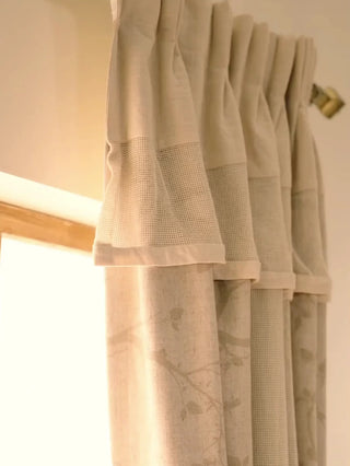 The Reed Rhythm readymade curtain in beige with a faint botanical print, shown in pleats on a brass rod and tied back beside a window