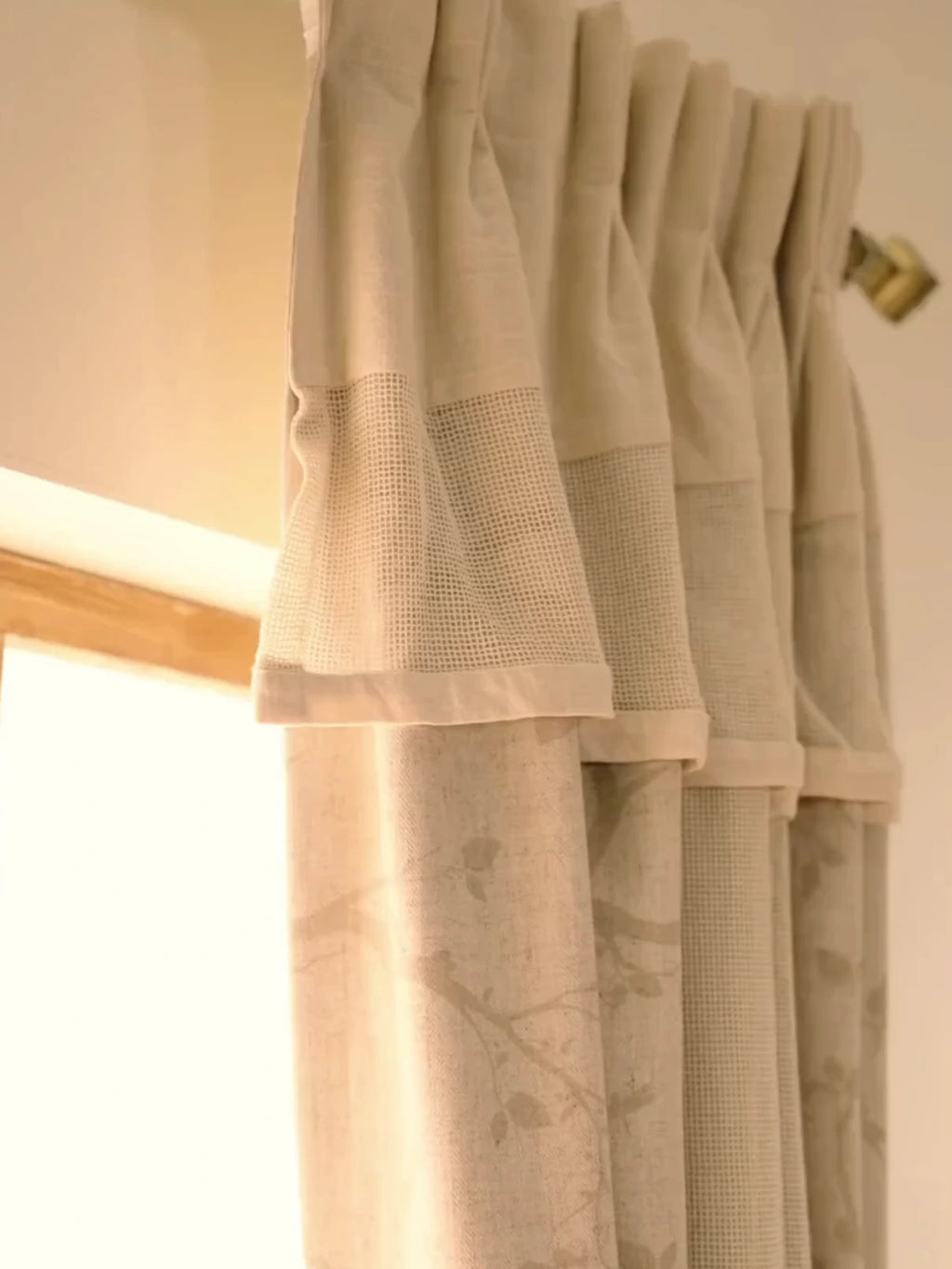 The Reed Rhythm readymade curtain in beige with a faint botanical print, shown in pleats on a brass rod and tied back beside a window