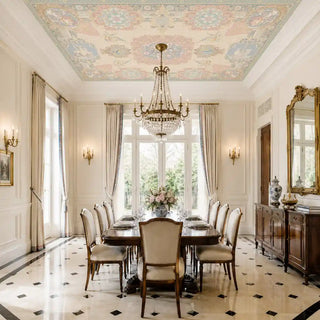 The Rahnuma ceiling mural in Dusty Rose & Gold installed above a formal dining room with a wood dining table set for a meal and a crystal chandelier. view-ceiling