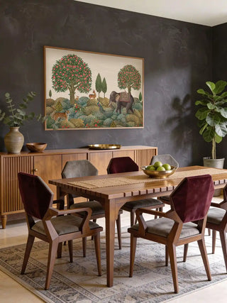 A grand dining room featuring the Rajasi Van forest mural above a wooden table with velvet chairs and a large fiddle leaf fig plant.