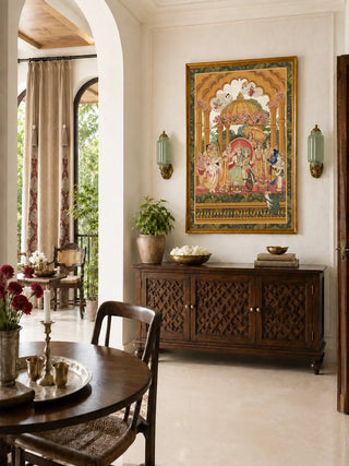 Ram Abhishek embroidered wall art in a gilt frame above a dark carved lattice sideboard with brass bowls and a potted plant in a sunlit dining room
