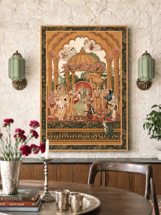 Ram Abhishek embroidered wall art in a slim gold frame on a textured white stone wall flanked by green glass sconces above a wooden sideboard in a dining room