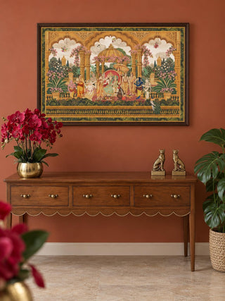 The Ram Abhishek embroidered wall art in a dark wood frame above a walnut console with brass knobs, red orchids and gilt leopard bookends.