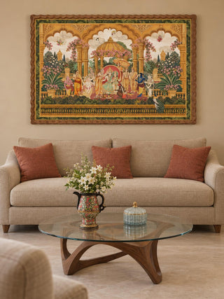 The Ram Abhishek embroidered wall art in a scalloped wood frame above an oatmeal sofa with terracotta cushions and a glass coffee table.