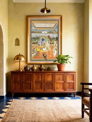 The Ram Darbar wall art in a gold frame styled above a large wooden console table with brass vessels and a green plant in a luxury foyer.