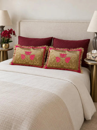A pair of Rangrez lumbar cushions on a cream bed before maroon pillows, gold elephants and a pink lotus on a gold ground with pink brocade borders.
