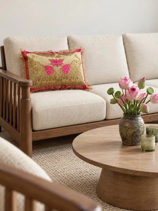 The Rangrez lumbar cushion on a cream sofa with a wooden frame, gold-embroidered elephants beside a pink lotus, edged in pink brocade and fringe.