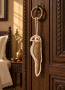 A hand-embroidered Ranjha charm in gold and ivory, shaped as a parrot with sequinned plumage, hung by rope from a brass ring door handle