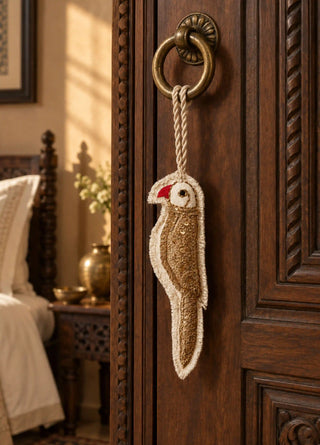 A hand-embroidered Ranjha charm in gold and ivory, shaped as a parrot with sequinned plumage, hung by rope from a brass ring door handle