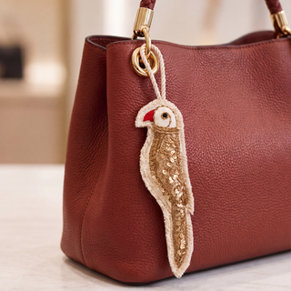 The Ranjha Parrot Hanging Charm in Ivory & Gold, clipped to a rust leather tote bag, showing the sequinned body and red beak.