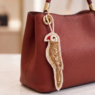 The Ranjha Parrot Hanging Charm in Ivory & Gold, clipped to a rust leather tote bag, showing the sequinned body and red beak.