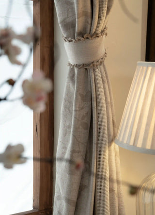 Close-up of the Reed Rhythm curtain in beige, gathered with a crochet-edged tieback, showing a tonal leaf print and open-weave mesh panel.