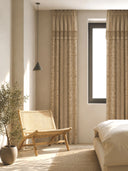 The Reed Rhythm beige pinch-pleat curtain panels styled beautifully in a minimalist organic bedroom beside a woven cane accent chair.