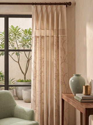 The Reed Rhythm Readymade neutral linen pinch-pleat curtain panel styled across a patio window next to a green armchair and wooden console table.