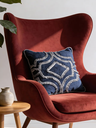 A luxury navy blue velvet cushion with a silver-grey fringed crest design styled on a rust-colored velvet wingback chair.