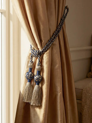 A luxury navy and gold braided curtain tieback with oversized tassels holding a champagne-colored silk drape.