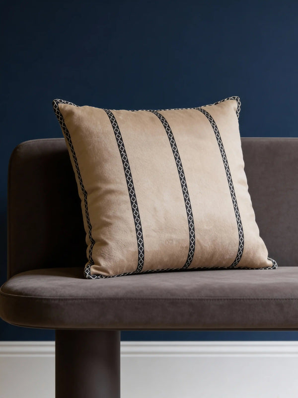 A luxury sandy-beige velvet cushion with black-and-white geometric embroidered vertical bands styled on a dark grey sofa against a navy blue wall.